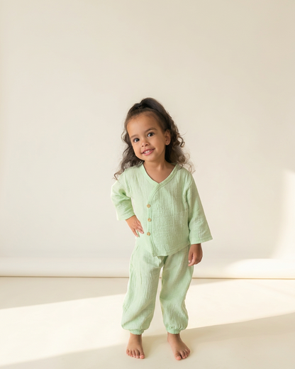 Meisje draagt een zachtgroen mousseline kimono-topje met bijpassende broek uit de zomercollectie, gefotografeerd tegen een neutrale studio-achtergrond – luchtige biologisch katoenen kinderkleding.